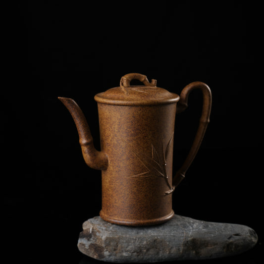 This shows a purple clay teapot with a brown body in the shape of a bamboo joint, a short spout, a curved handle, and bamboo leaf carvings on the body. It is placed on an irregularly shaped stone base.