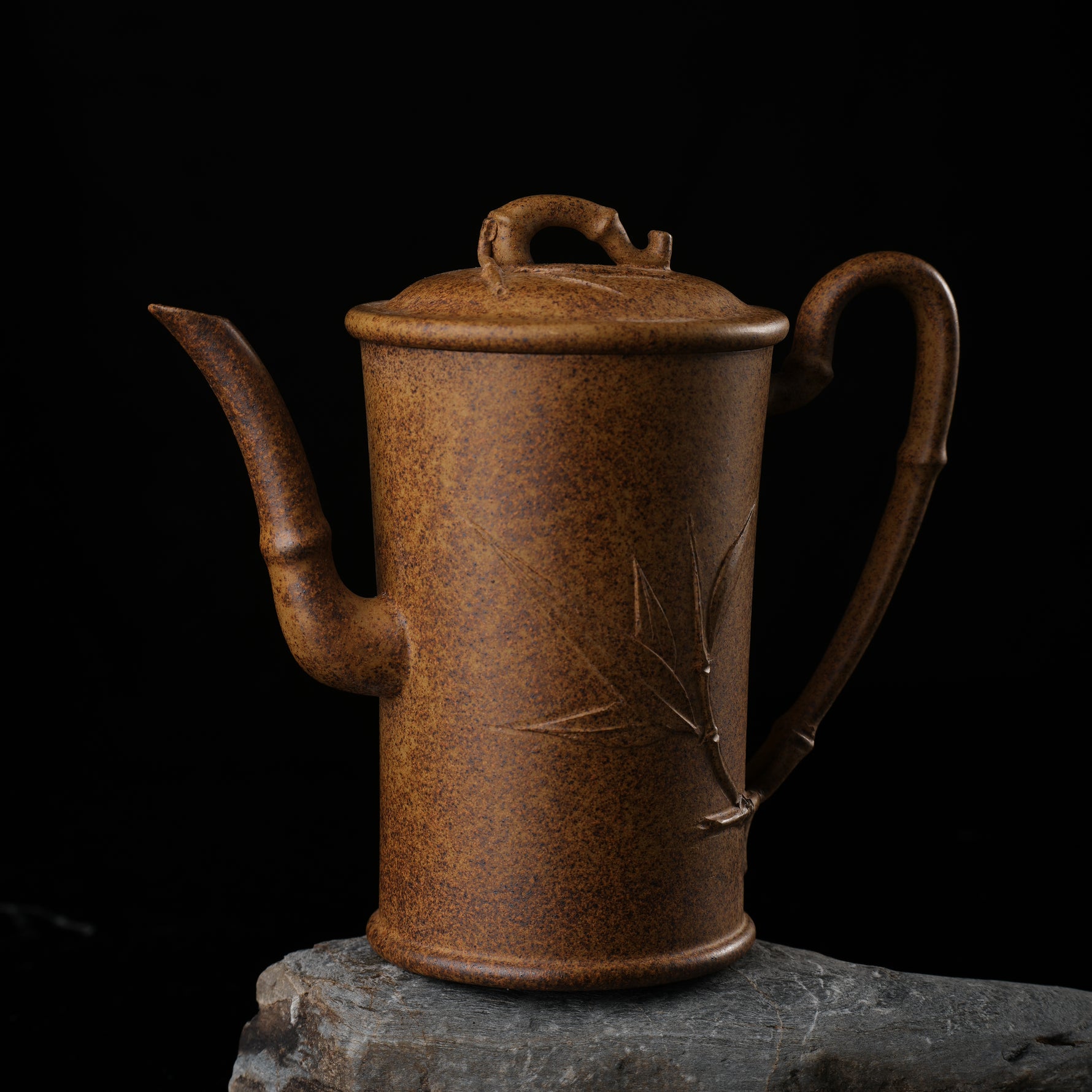 This shows a purple clay teapot with a brown body in the shape of a bamboo joint, a short spout, a curved handle, and bamboo leaf carvings on the body. It is placed on an irregularly shaped stone base.