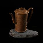 This shows a purple clay teapot with a brown body in the shape of a bamboo joint, a short spout, a curved handle, and bamboo leaf carvings on the body. It is placed on an irregularly shaped stone base.