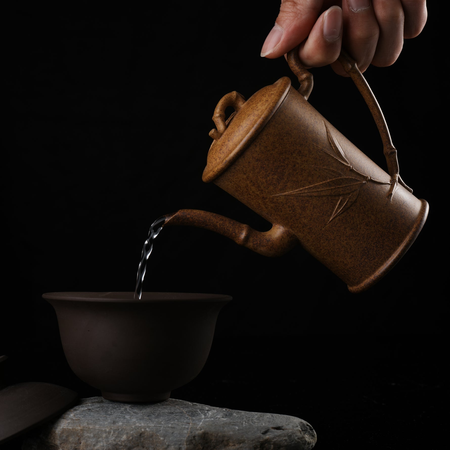 This shows a purple clay teapot with a brown body in the shape of a bamboo joint, a short spout, a curved handle, and bamboo leaf carvings on the body. It is placed on an irregularly shaped stone base.