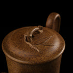 This shows a purple clay teapot with a brown body in the shape of a bamboo joint, a short spout, a curved handle, and bamboo leaf carvings on the body. It is placed on an irregularly shaped stone base.