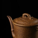 This shows a purple clay teapot with a brown body in the shape of a bamboo joint, a short spout, a curved handle, and bamboo leaf carvings on the body. It is placed on an irregularly shaped stone base.