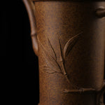 This shows a purple clay teapot with a brown body in the shape of a bamboo joint, a short spout, a curved handle, and bamboo leaf carvings on the body. It is placed on an irregularly shaped stone base.