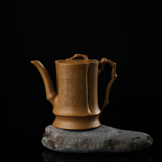 This shows a purple clay teapot with a warm yellow body, engraved with words, a short spout, a well-curved handle, and a tightly fitting lid. The purple clay teapot is placed on a gray irregular-shaped base.