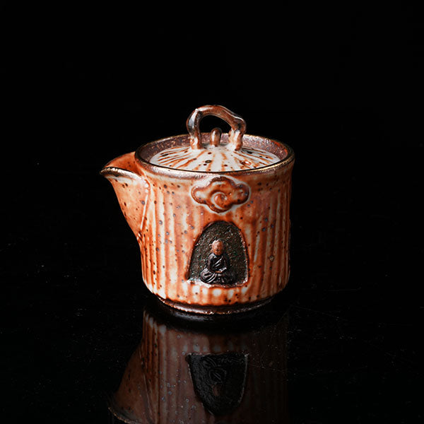 A traditional ceramic teapot with a rustic, speckled brown-orange glaze. It features a lid with a handle, a meditating figure relief, and intricate textural designs. Ideal for tea ceremonies, embodying Asian art and mindfulness. Handcrafted, showcasing cultural heritage and artistic detail. Perfect for collectors of unique teaware, highlighting traditional craftsmanship and spiritual motifs. 