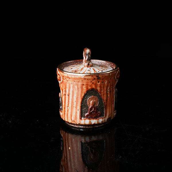 A traditional ceramic teapot with a rustic, speckled brown-orange glaze. It features a lid with a handle, a meditating figure relief, and intricate textural designs. Ideal for tea ceremonies, embodying Asian art and mindfulness. Handcrafted, showcasing cultural heritage and artistic detail. Perfect for collectors of unique teaware, highlighting traditional craftsmanship and spiritual motifs. 